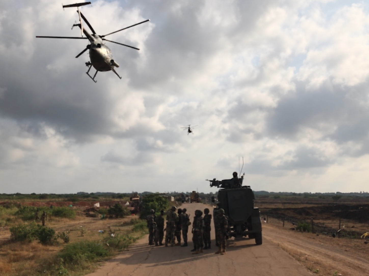 Helicopters above personnel and a military vehicle on a rural road